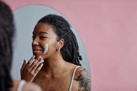 Candid portrait of young black woman with acne scars using face cream looking in mirror reflection