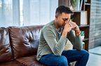 A man sits on the couch with his elbows on his knees, hands folded to his face with his eyes closed