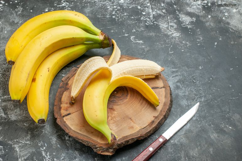 A bundle of fresh bananas and a peeled banana on a wooden cutting board with a knife