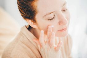 Young woman in bathrobe applying moisturizing cream on face.