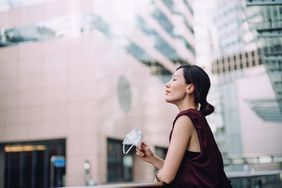 Young Asian woman taking off protective face mask, setting herself free and feeling relieved. Head up with her eyes closed. Taking a break from busy and stressful work life in downtown district in the city