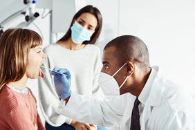 mom and daughter at the doctor's office. doctor is wearing a mask and checking the daughter's mouth