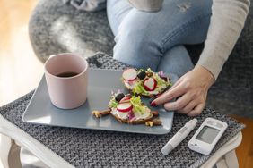A woman eats her snack with her glucometer next to her on the table.