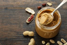 jar of peanut butter with spoon of peanut butter with peanuts sitting next to it