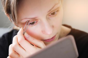 A young woman is plucking her eyebrows with tweezers in the mirror