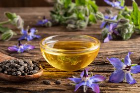 Small glass bowl of borage oil (yellow-amber in color) on a wood table, surrounded by seeds and purple flowers from the borage plant 