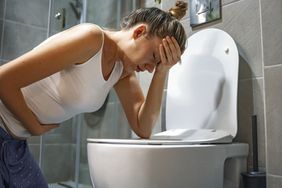 Female with hand on head and stomach leaning over toilet with look of pain on face