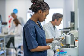 Female technician in late 20s examining blood sample as she organizes test tubes for centrifuge in clinical analysis laboratory.