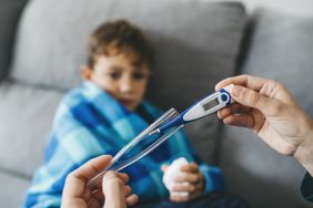 Sick boy with Kawasaki disease sitting on the couch and his mother's hands preparing to place the thermometer in the armpit