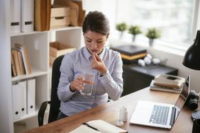 Female taking medicine while sitting at desk with computer and glass of water