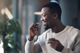 Young black man wearing a cream sweater is taking a supplement and holding a glass of water. 