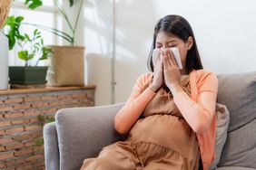 Young Asian pregnant woman blowing nose with a tissue while sitting on a couch