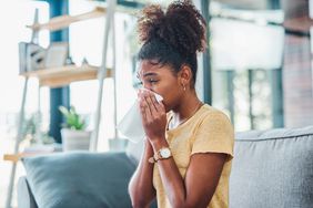 Young black woman wearing a yellow t-shirt sitting on her couch and blowing her nose with a tissue
