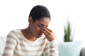 Young black woman wearing striped sweating and sitting on her couch, holding her nose in pain from sinus pressure
