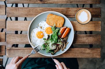 Person eating a plate of food: eggs, hashbrowns, sausage, greens; on a wooden table with a latte.