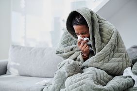 person wrapped in blanket sitting on couch sneezing into a tissue