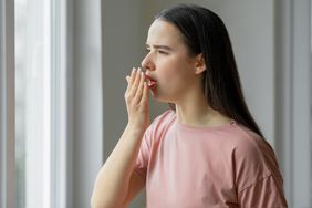 White woman with brown hair wearing light pink tshirt holds her hand in front of her mouth, smelling her breath