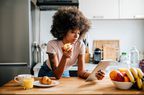 woman with afro hairstyle is leaning over a kitchen counter holding an apple and looking at a tablet.