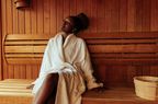 A young Black woman is sitting on the bench in the sauna wearing her bathrobe.