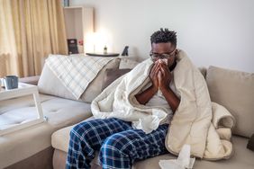 Black man sitting on couch wrapped in blanket blowing nose