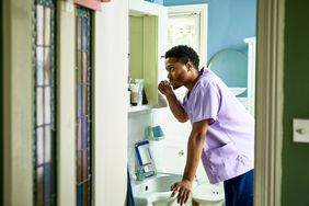 Young Black man looking in mirror using toothbrush, leaning on edge of basin