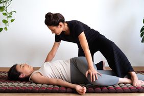 Female massage therapist is applying Thai massage technique to young woman. The therapist crouches over the client laying down and holds her knee to the side in a spinal twist. 