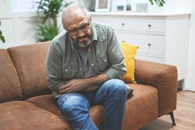 Older man with beard sitting on couch holding stomach with expression of pain on face
