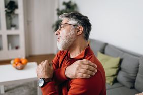 older man sitting on living room sofa holding his painful shoulder