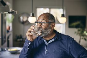 Older black man wearing glasses and blue shirt drinking water in his kitchen