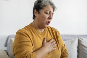 Middle-aged woman with short gray hair wearing a yellow sweater touching her palm to her chest with a pained expression
