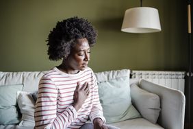 Black woman with hand on chest, wearing a white and pink striped shirt, sitting on couch