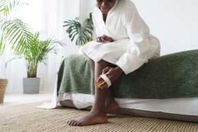 Black female massaging leg with dry brush sitting on bed