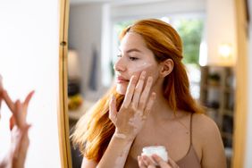 Young white woman with red hair and vitiligo applying cream to her face in a mirror