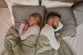 Female/male couple in bed lying back to back, awake. Bedding is green, white, and grey. Both people are wearing long sleeve white t shirts 