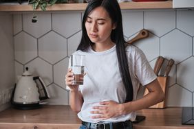 Sick young Asian woman standing in kitchen holding glass of water suffering from stomach ache, 