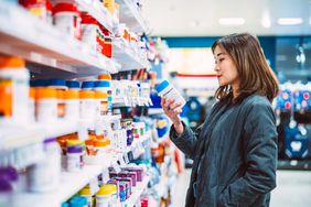 Asian woman reading the ingredient label while shopping for multivitamin and health supplements on shelf in supermarket.
