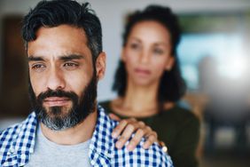 Man looking distressed, woman in the background blurry touching his shoulder