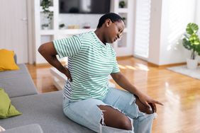 Young black woman wearing jeans and a striped tshirt is sitting on a couch and holding her lower back in pain