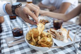 man picking up a chip out of a bowl of chips and olives on a table with a checkered tablecloth