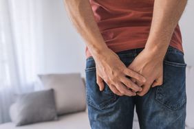 Close up of a man in jeans and a red t-shirt in his living room. The man is holding the back of his pants after experiencing a shart