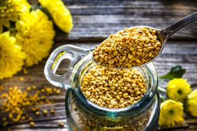 A metal spoon scoops bee pollen (small yellow nuggets) out of a glass jar. The jar sits on a rustic wooden table with yellow flowers in the background.