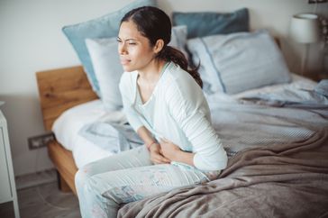 A woman clutches her stomach in pain as she sits on the side of her bed