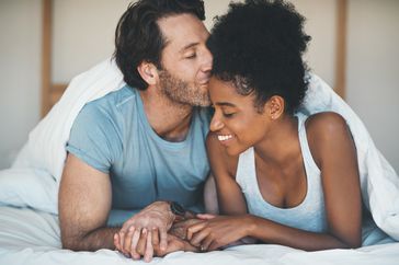 a young couple in bed together: a white man with scruff wearing a blue tshirt kisses a black woman wearing a gray tank-top on the forehead. They are holding hands