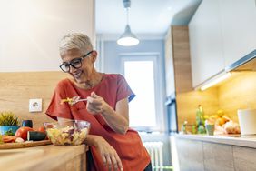 Senior woman standing in the kitchen and relaxing at home while eating a small green salad.