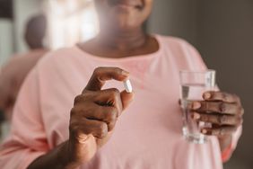 Closeup of a Black woman standing in her bathroom wearing a pink shirt, holding a white supplement pill in one hand and a glass of water in the other