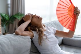 A woman sits on her couch, hot, as she fans herself for some relief