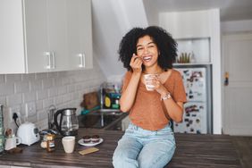 Young woman with curly hair wearing jeans and an orange tshirt is sitting on a kitchen countertop eating yogurt