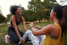 A black woman in a sports bra with her hair pulled back training a client through a core workout. The client has on a yellow shirt and newspaper print pants.