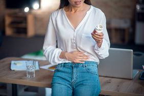 Young woman wearing jeans and a white blouse standing in an office setting, holding her stomach and looking at a supplement pill bottle.