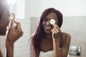 Smiling African American woman standing at bathroom and putting fresh cucumber on her eye.